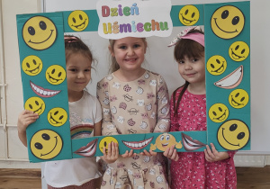 Dziewczynki pozują do zdjęcia stojąc za ramką ozdobioną uśmiechami.