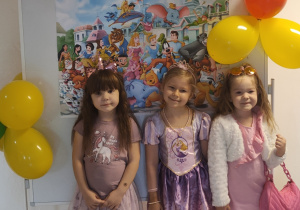 Trzy dziewczynki w bajkowych strojach pozują do zdjęcia na tle dekoracji z okazji Dnia Postaci z Bajek.