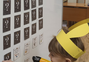 Chłopiec stojąc przed tablicą magnetyczną odkrywa kolejną kartę z zagadką o wybranej postaci bajkowej.