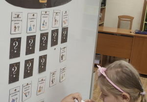 Dziewczynka stojąc przed tablicą magnetyczną trzyma w rękach kartkę z zagadką o wybranej postaci bajkowej.