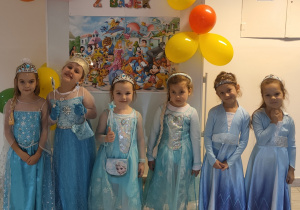 Grupa dziewczynek w bajkowych strojach pozuje do zdjęcia na tle dekoracji z okazji Dnia Postaci z Bajek.