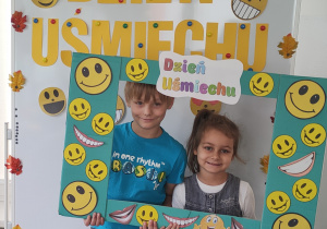 Chłopiec i dziewczynka pozują do zdjęcia stojąc za ramką ozdobioną uśmiechami.