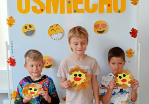 Chłopcy pozują do zdjęcia z własnoręcznie wykonanymi uśmiechami z talerzyków papierowych. Talerzyk papierowy jest w kolorze żółtym, na nim jest narysowana uśmiechnięta buźka a wokół przyklejone są listki.