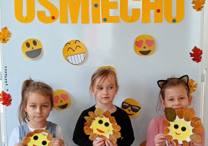 Dziewczynki pozują do zdjęcia z własnoręcznie wykonanymi uśmiechami z talerzyków papierowych. Talerzyk papierowy jest w kolorze żółtym, na nim jest narysowana uśmiechnięta buźka a wokół przyklejone są listki.