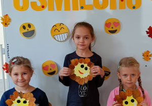Dziewczynki pozują do zdjęcia z własnoręcznie wykonanymi uśmiechami z talerzyków papierowych. Talerzyk papierowy jest w kolorze żółtym, na nim jest narysowana uśmiechnięta buźka a wokół przyklejone są listki.