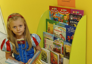 Widok na dziewczynkę siedzącą na krześle z otwartą książką przy biblioteczce przedszkolnej.