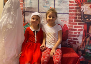 Dwie dziewczynki siedząc na ławeczce pozują do zdjęcia. W tle dekoracja świąteczna.