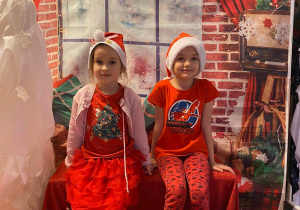 Dwie dziewczynki siedząc na ławeczce pozują do zdjęcia. W tle dekoracja świąteczna.