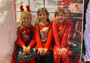 Trzy dziewczynki siedząc na ławeczce pozują do zdjęcia. W tle dekoracja świąteczna.