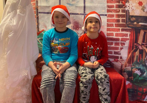 Dwóch chłopców siedząc na ławeczce pozuje do zdjęcia. W tle dekoracja świąteczna.