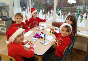 Widok na siedzące przy stoliku dzieci, które ozdabiają papierowy szalik kredkami oraz kolorową bibułą.