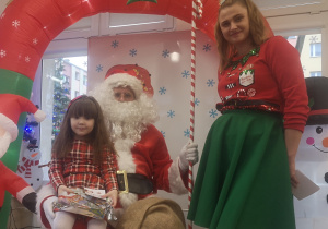 Dziewczynka siedzi na kolanach św. Mikołaja, trzymając w dłoniach prezent. Obok stoi pani Elf.