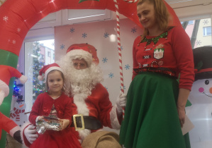 Dziewczynka siedzi na kolanach św. Mikołaja, trzymając w dłoniach prezent. Obok stoi pani Elf.