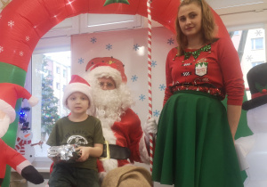 Chłopiec siedzi na kolanach św. Mikołaja, trzymając w dłoniach prezent. Obok stoi pani Elf.
