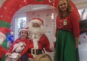 Chłopiec siedzi na kolanach św. Mikołaja, trzymając w dłoniach prezent. Obok stoi pani Elf.