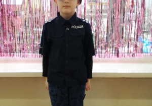 Chłopiec w stroju policjanta pozuje do zdjęcia. W tle kurtyna dekoracyjna w kolorze różowym.