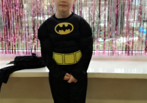 Chłopiec w stroju Batmana pozuje do zdjęcia. W tle kurtyna dekoracyjna w kolorze różowym.