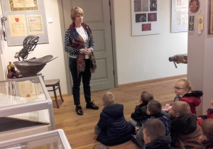 Dzieci słuchają jak pracownik muzeum opowiada o eksponatach.
