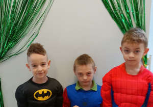 Chłopcy pozują do zdjęcia w strojach karnawałowych. W tle tablica magnetyczna z napisem "Karnawał" udekorowana kurtyną dekoracyjną w kolorze zielonym.