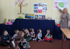 Widok na siedzące dzieci, które spoglądają na symbole narodowe prezentowane przez Panią z biblioteki.