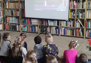 Widok na siedzące dzieci przed ekranem projektora, które słuchają piosenki o Polsce.