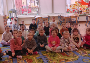 Grupa dzieci siedzi na dywanie i uważnie słuchają pani z AleKarmel.