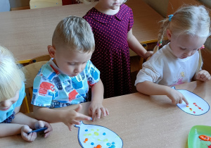 Dzieci siedzą przy stoliku. Stemplują palcami kropki na kartonie w kształcie balonika.