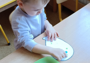 Chłopiec siedzi przy stoliku. Stempluje palcami kropki na kartonie w kształcie balonika.