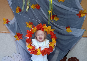 Dziecko pozuje do zdjęcia trzymając w rękach obręcz z jesiennymi liśćmi.