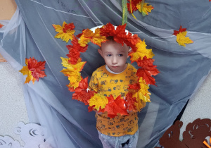 Dziecko pozuje do zdjęcia trzymając w rękach obręcz z jesiennymi liśćmi.