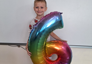 Chłopiec pozuje do zdjęcia, w rękach trzyma balona w kształcie cyfry 6.