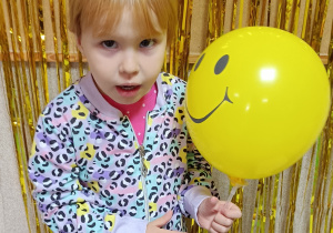 Dziewczynka pozuje do zdjęcia, w dłoni trzyma żółty balon, na którym znajduje się uśmiech.