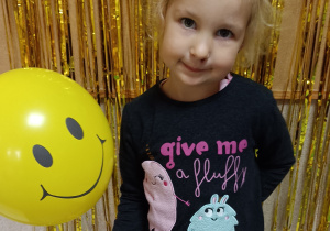 Dziewczynka pozuje do zdjęcia, w dłoni trzyma żółty balon, na którym znajduje się uśmiech.