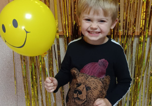Chłopiec pozuje do zdjęcia, w dłoni trzyma żółty balon, na którym znajduje się uśmiech.