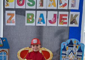Chłopiec w stroju strażaka pozuje do zdjęcia. W tle znajduje się dekoracja z okazji Dnia Postaci z Bajek.