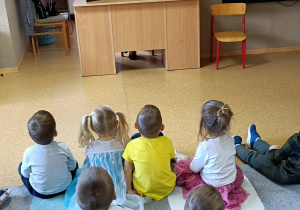 Widok na siedzące dzieci, które z uwagą oglądają bajkę "Trzy świnki" prezentowaną w formie teatrzyku kamishibai przez Panią z biblioteki.