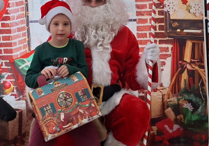Dziecko siedzi na kolanach u Świętego Mikołaja. W tle świąteczna dekoracja.