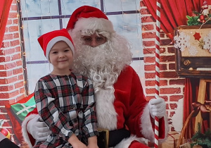 Dziecko siedzi na kolanach u Świętego Mikołaja. W tle świąteczna dekoracja.