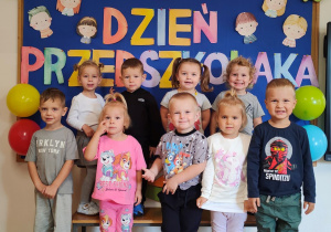 Dzieci pozują do zdjęcia grupowego. W tle znajduje się dekoracja z okazji Dnia Przedszkolaka.