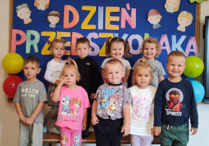 Dzieci pozują do zdjęcia grupowego. W tle znajduje się dekoracja z okazji Dnia Przedszkolaka.