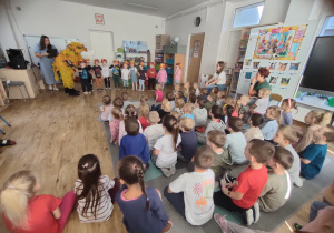 Widok na dzieci, które siedzą i z zainteresowaniem obserwują maluszki prezentujące jesienną piosenkę w kolorowych kapeluszach.
