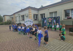 Widok na tańczące dzieci, które poruszają kolorowymi chustkami w rytm muzyki, na tle przygotowanej dekoracji.