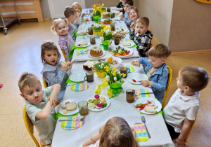 Widok na dzieci siedzące przy pięknie udekorowanym wielkanocnym stole, które wspólnie spożywają przygotowane świąteczne śniadanie.