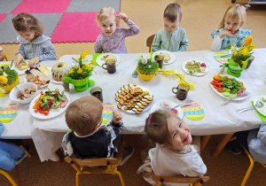Widok na dzieci siedzące przy pięknie udekorowanym wielkanocnym stole, które wspólnie spożywają przygotowane świąteczne śniadanie.