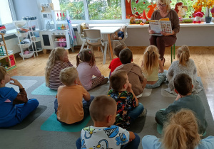 Widok na dzieci siedzące na dywanie, uczestniczące w zajęciach prowadzonych przez Panią z biblioteki.