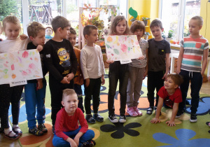 Dzieci stojące w półkolu prezentują prace plastyczne na temat: warzywa, owoce.
