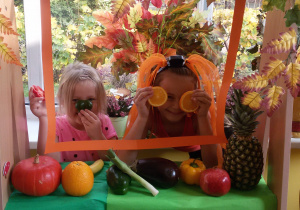 Dwie dziewczynki prezentują w okienku śmieszne minki, wykorzystując plastry i kawałki owoców.