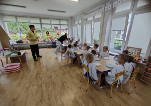 Widok na dzieci siedzące przy stole w stroju chemika, które z uwaga słuchają Pana.