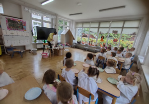 Widok na dzieci siedzące przy stole w stroju chemika, które obserwują eksperyment wykonywany przez Pana.