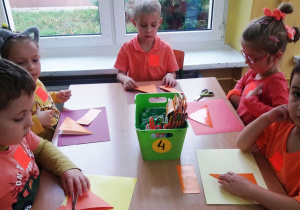 Troje dzieci siedzi przy stoliku, przed nimi leżą kolorowe kartki a na nich pomarańczowe kwadraty.